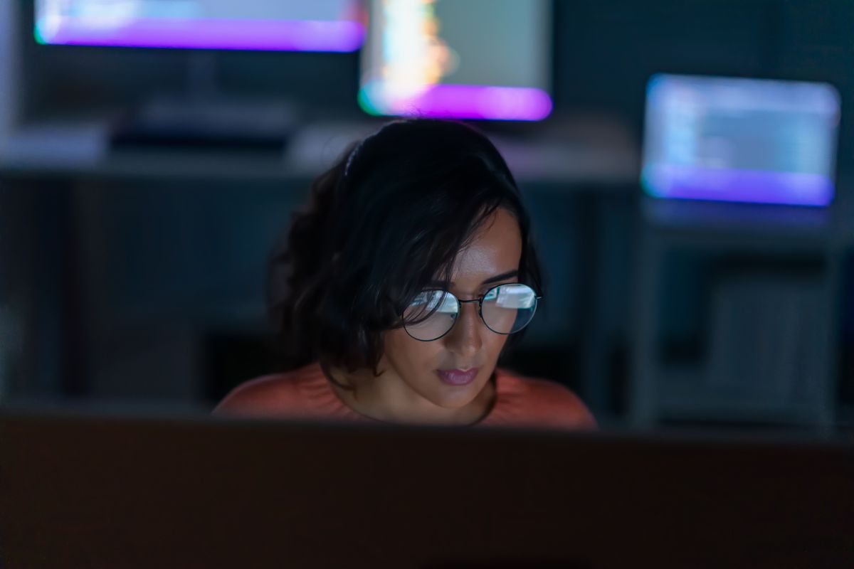 Female Java developer working at a computer station in low light with screen reflecting on glasses.