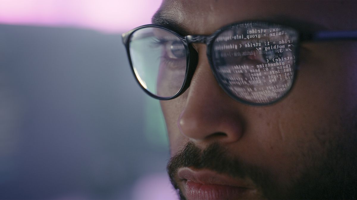Software developer working at computer screen conducting prompt engineering tasks with binary code and screen reflecting in glasses.