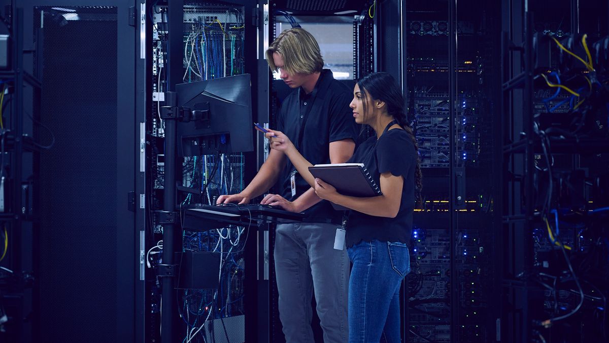 Data center outages concept image showing two technicians troubleshooting issues on a laptop in a server room.